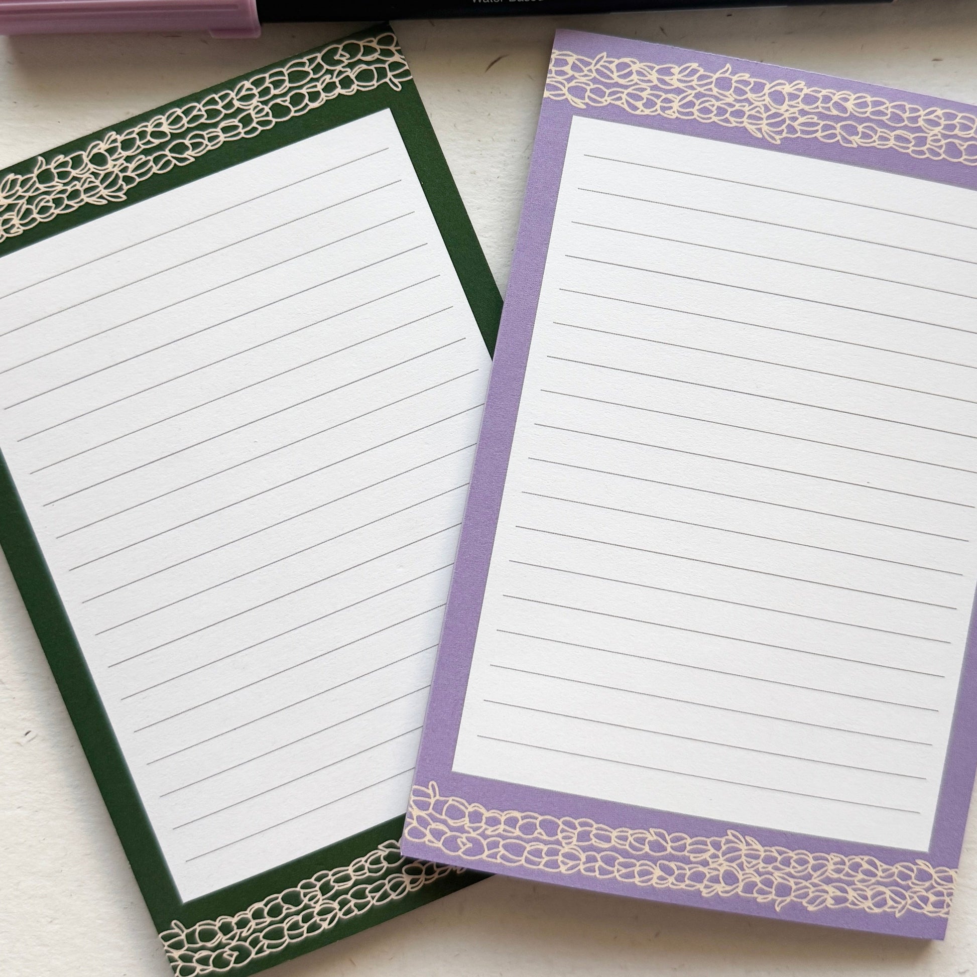 Two notepads with decorative borders and three pens on a white surface