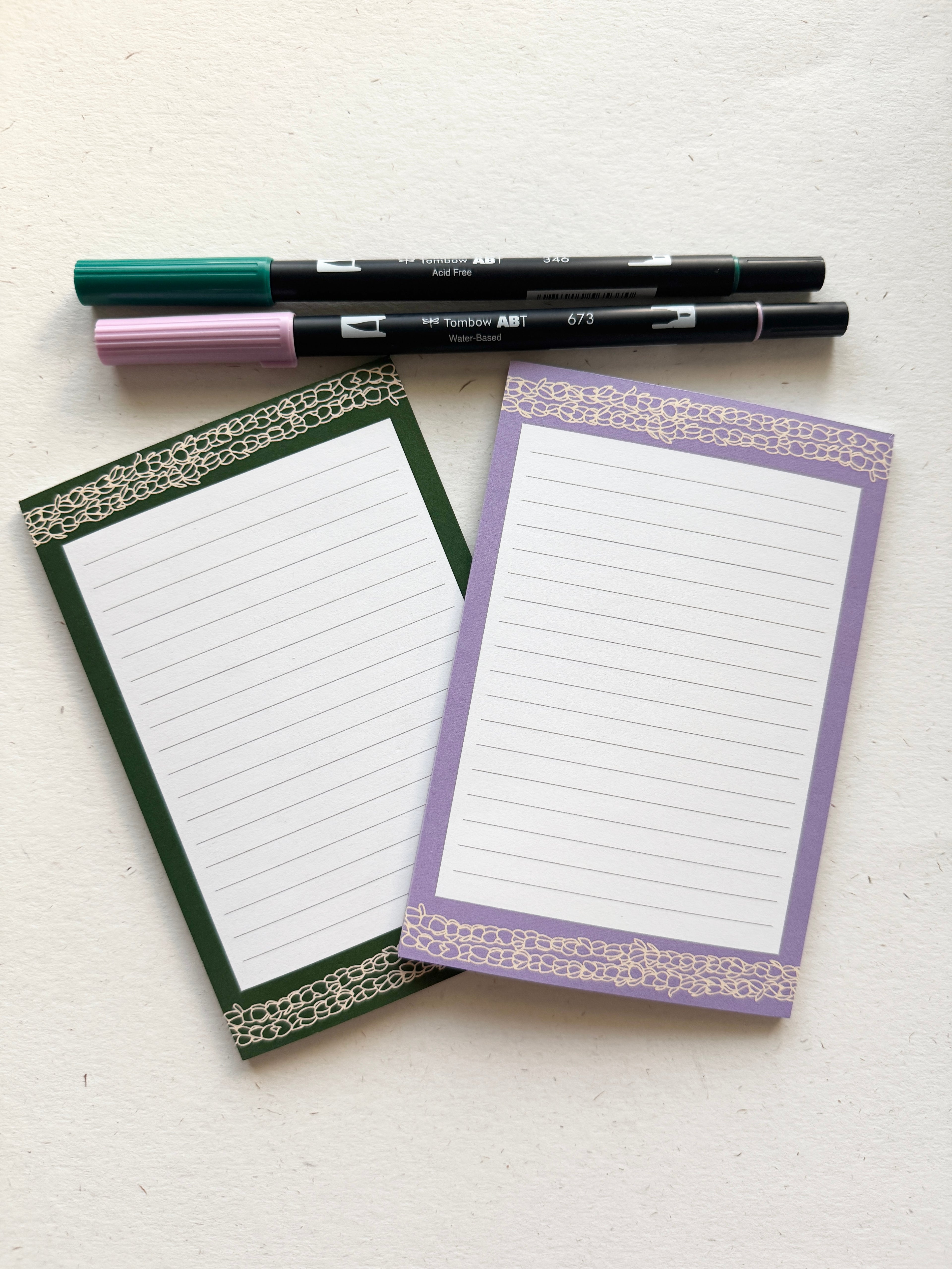 Two notepads with decorative borders and three pens on a white surface