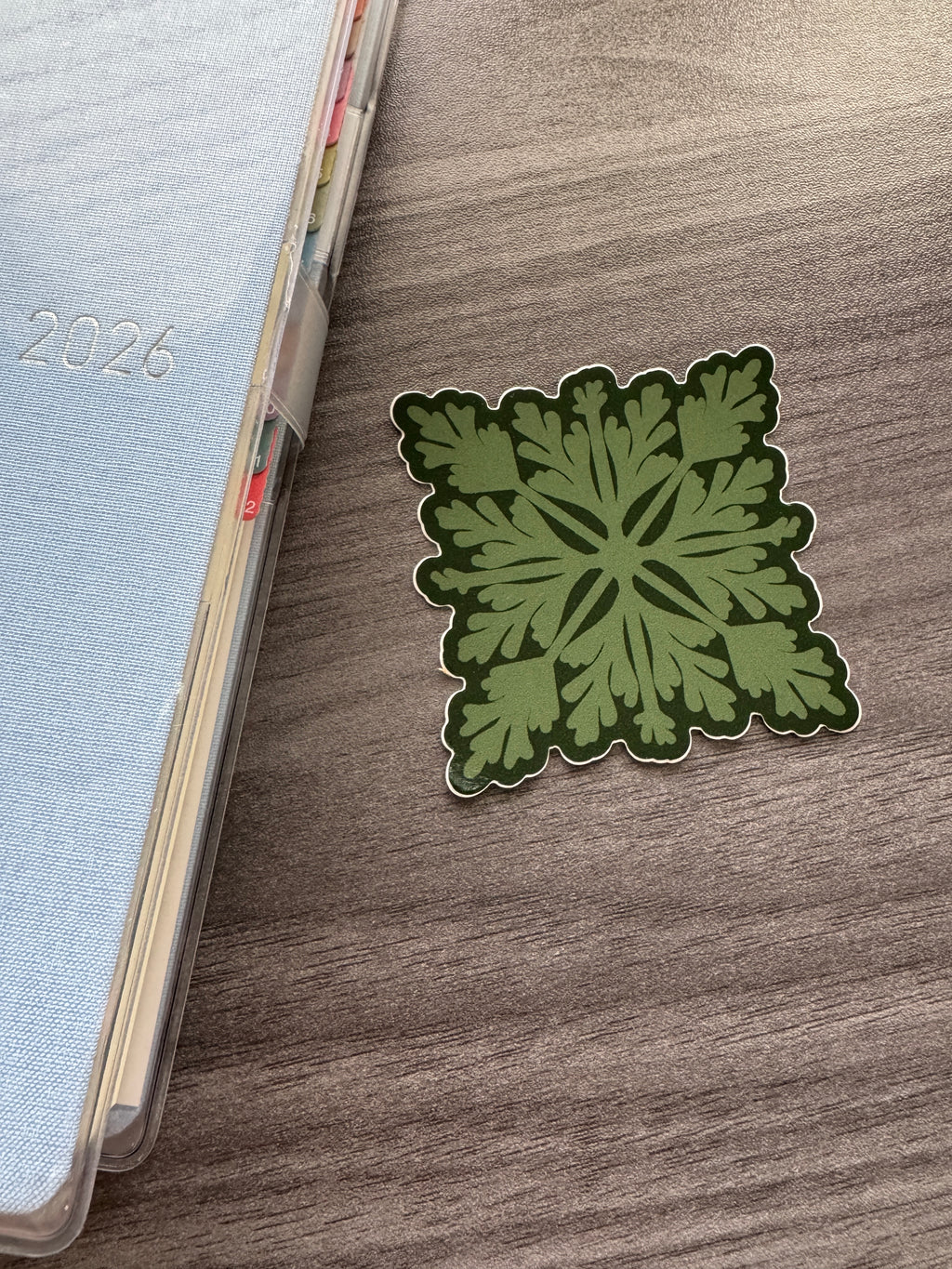 A square green pala'a quilt sticker with a dark green border and white outline, placed on a brown wood-grain tabletop next to a light blue planner.