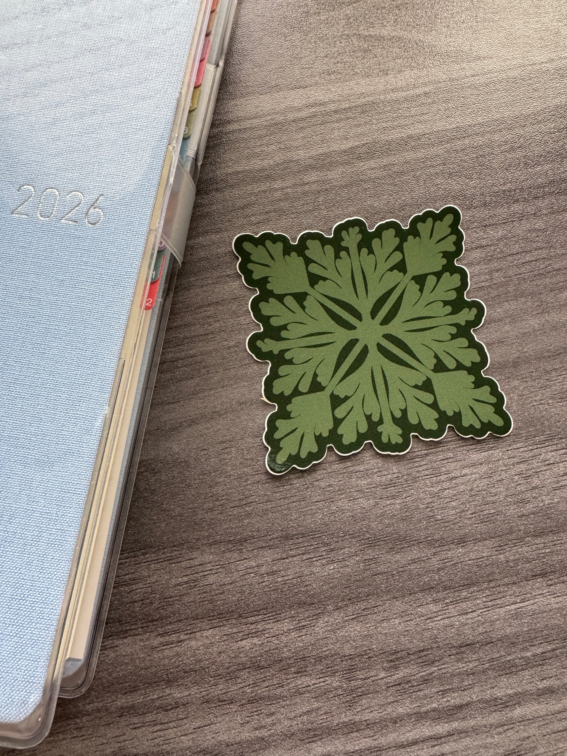 A square green pala'a quilt sticker with a dark green border and white outline, placed on a brown wood-grain tabletop next to a light blue planner.