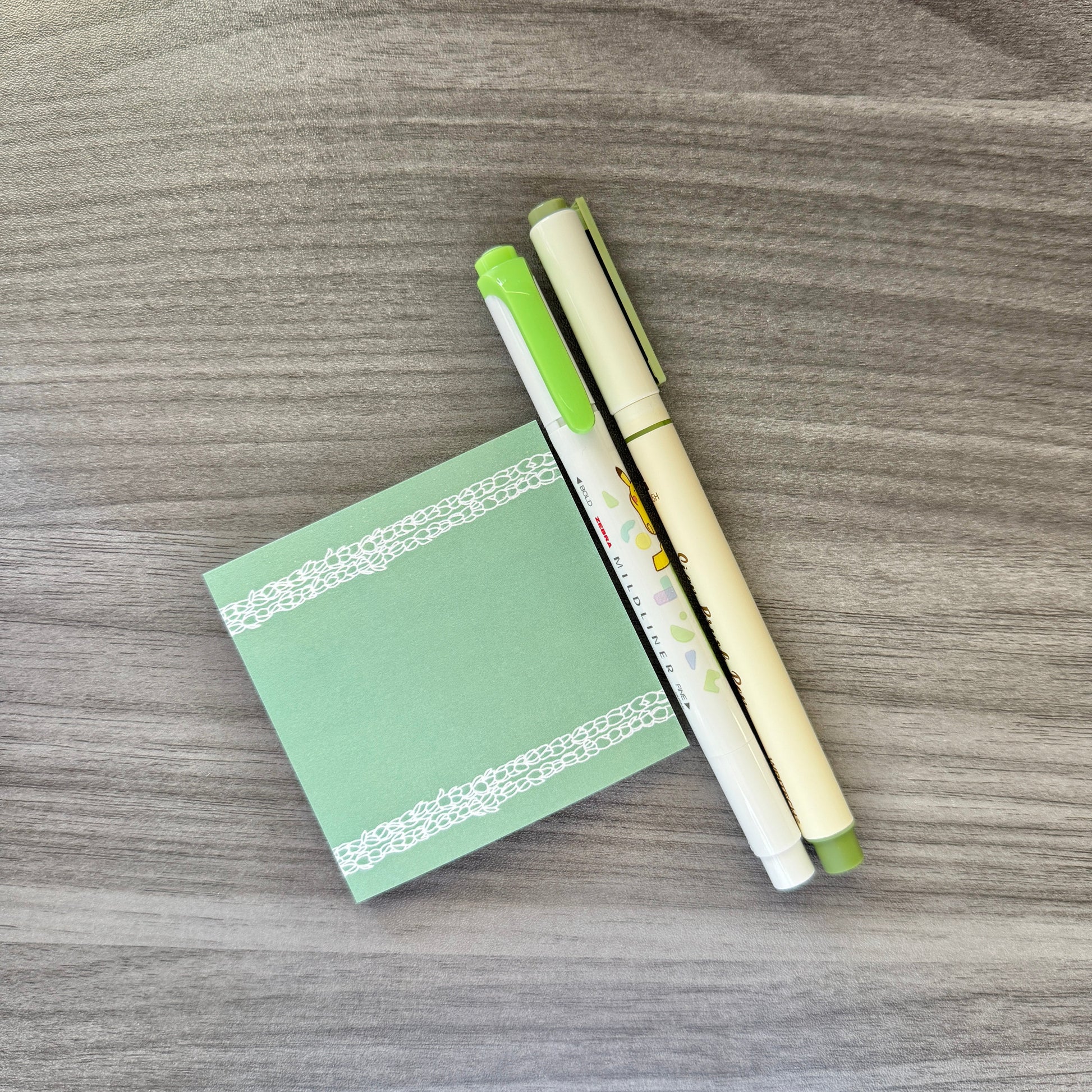 A small mint green sticky note pad with white dotted borders, placed on a gray wood-grain surface next to two cream-colored pens with green accents.