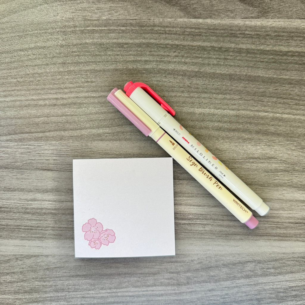 Two Sakura Pigma pencil markers with pink and cream barrels, placed diagonally next to a small white notepad with a pink cherry blossom graphic, all on a gray wood-grain surface.