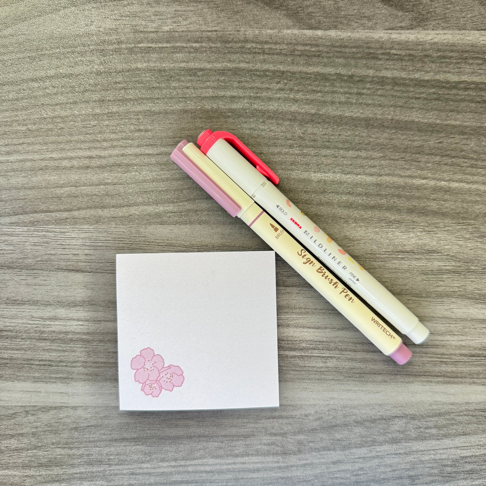 Two Sakura Pigma pencil markers with pink and cream barrels, placed diagonally next to a small white notepad with a pink cherry blossom graphic, all on a gray wood-grain surface.